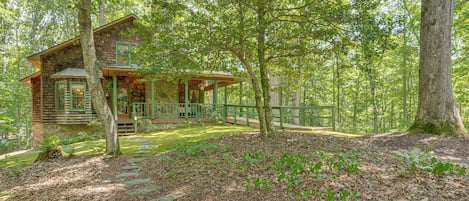 A cabin with a stone foundation and wooden siding, surrounded by a forest with a stone pathway leading to the entrance.