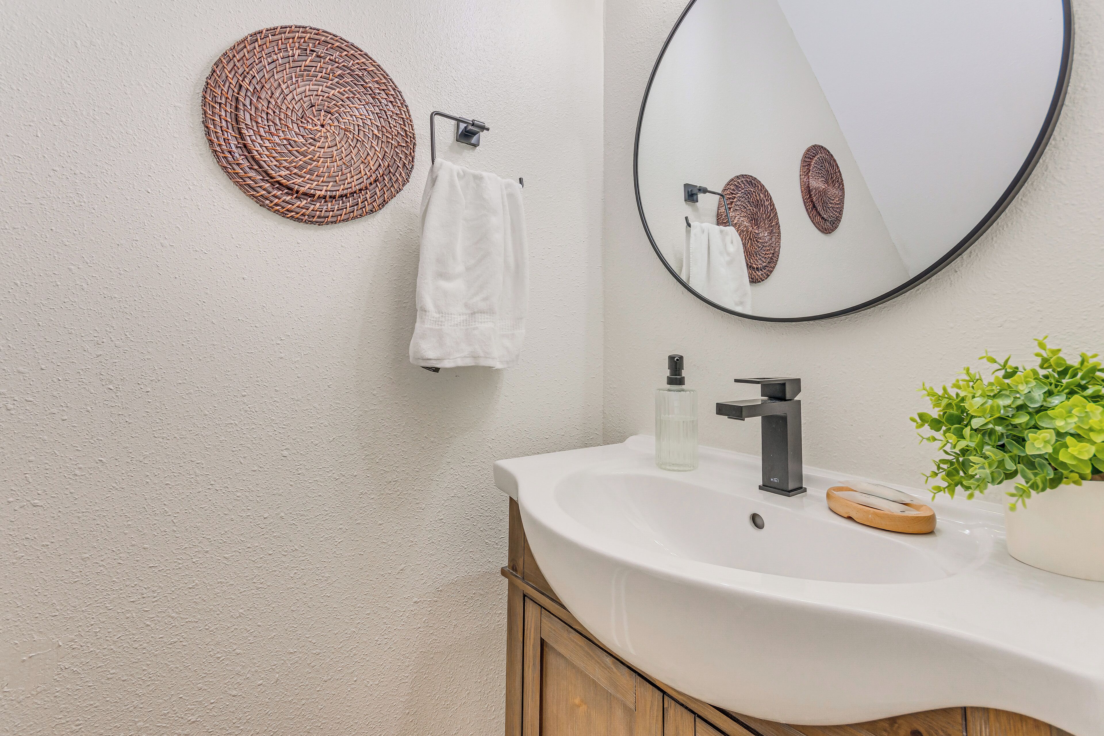 A bathroom with a white sink, a round mirror, a woven wall decoration, a white towel, and a green plant.