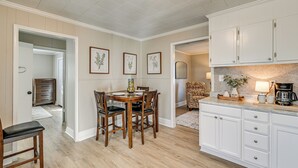 Un coin repas avec une table et des chaises en bois, une cuisine avec des meubles blancs et, au mur, des reproductions encadrées de gravures botaniques.