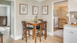 Un coin repas avec une table et des chaises en bois, des reproductions botaniques encadrées accrochées au mur, et un espace cuisine avec une chaise à motifs et des placards blancs.