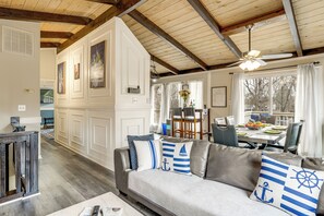Een woonkamer met een grijze bank, houten balken in het plafond en een eethoek met een tafel en stoelen.