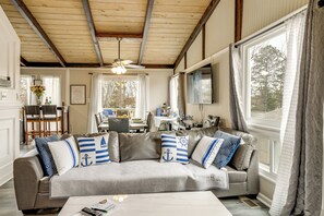 Een woonkamer met een grijze bank voorzien van kussens met nautische motieven, een houten plafond en een eethoek op de achtergrond.