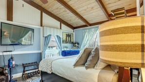 Une chambre à coucher avec un lit, un téléviseur fixé au mur, une fenêtre avec des rideaux, une petite table avec une lampe et un tapis à motifs.