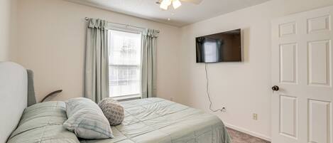 Un dormitorio con cama, ventilador de techo, ventana con cortinas, televisor de pantalla plana montado en la pared y una puerta.