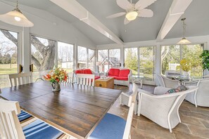 Une véranda spacieuse, dotée d'une grande table en bois, de meubles en osier blancs et de larges fenêtres offrant une vue sur l'extérieur.