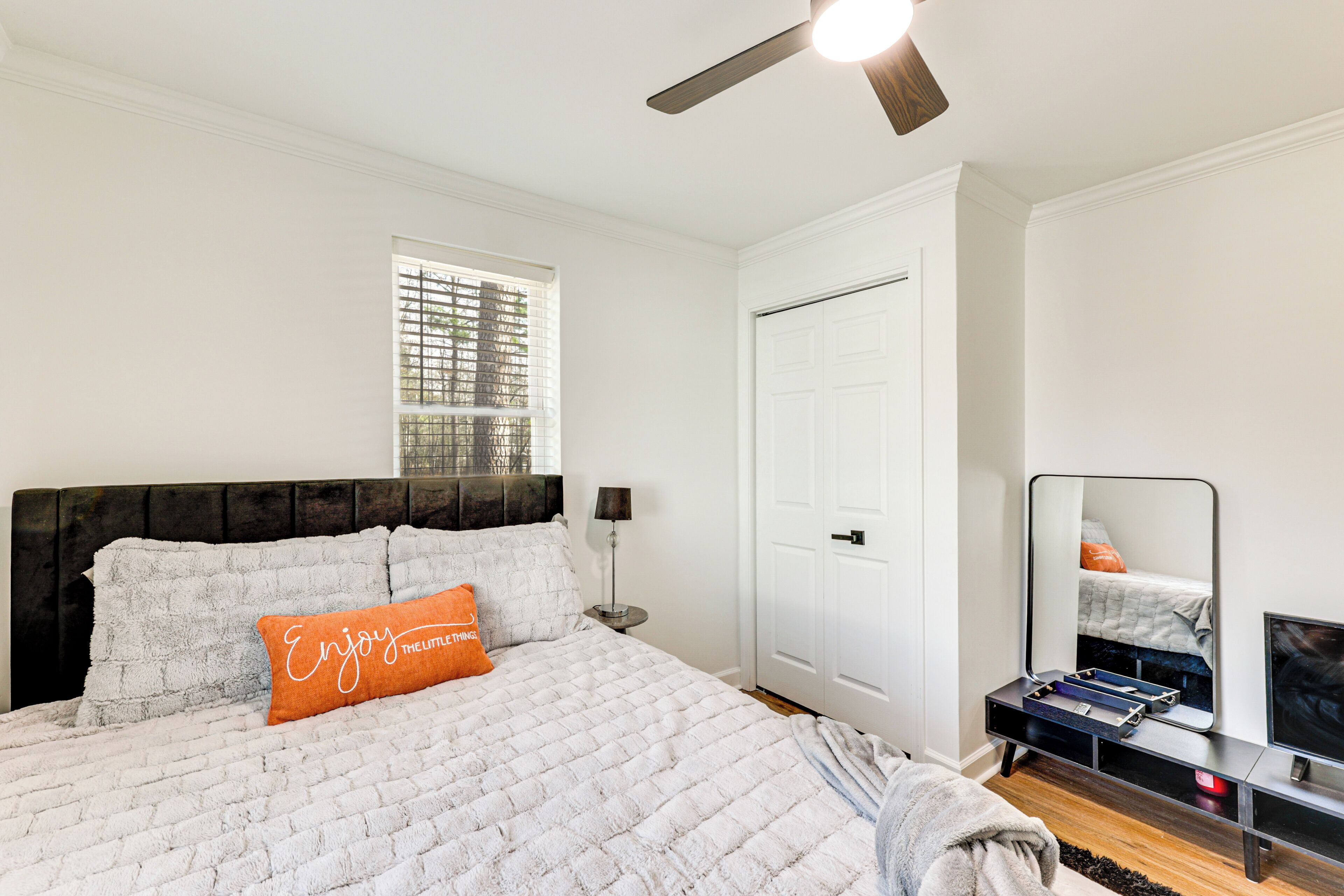 A bedroom with a bed, a ceiling fan, a window, a door, and a TV stand.