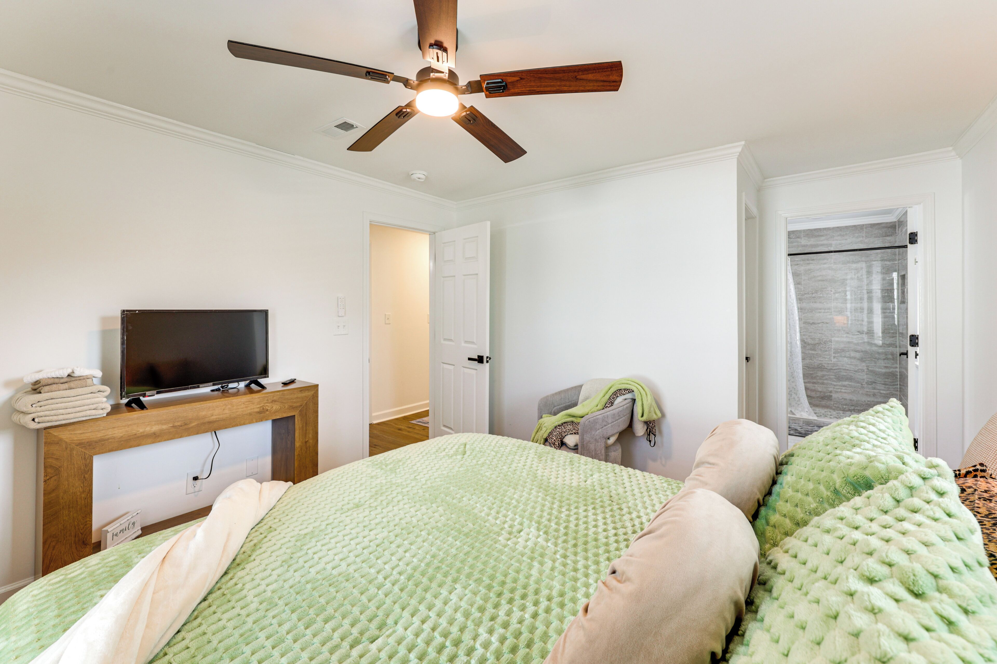 A bedroom with a bed, a TV on a wooden stand, a ceiling fan, and a door leading to another room.