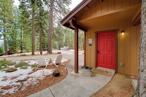 A wooden cabin with a red door, two Adirondack chairs outside, and a snow-covered ground.