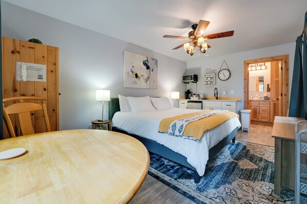 A hotel room with a bed, a wooden table, a ceiling fan, a microwave, and a clock on the wall.