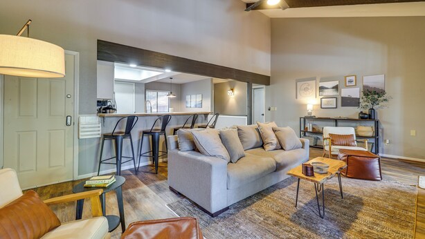 A spacious living room with a sofa, coffee table, and chairs. A kitchen area is visible in the background with bar stools.