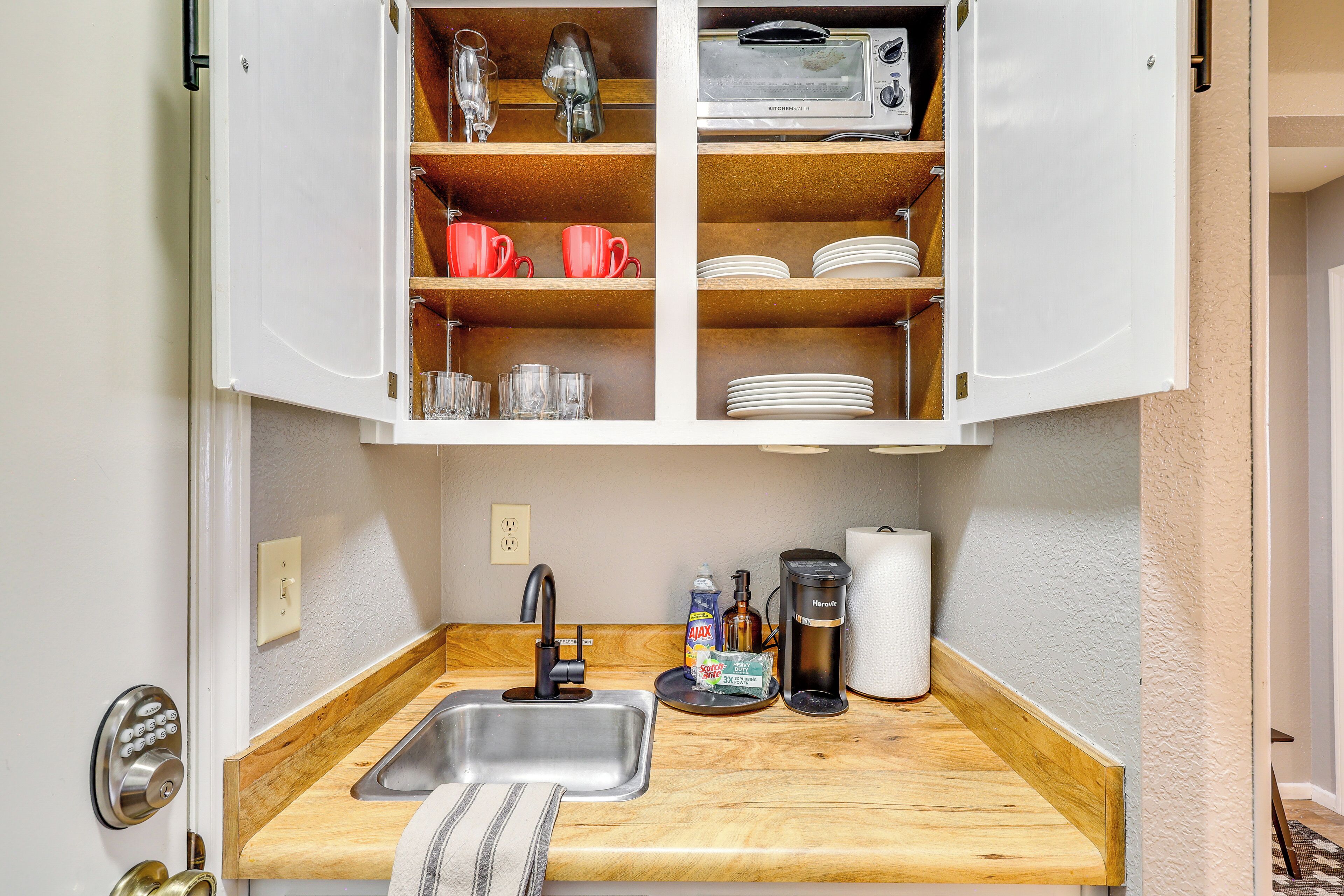 A compact kitchen with a built-in sink, wooden countertop, and open cabinets containing dishes, glasses, and a toaster.