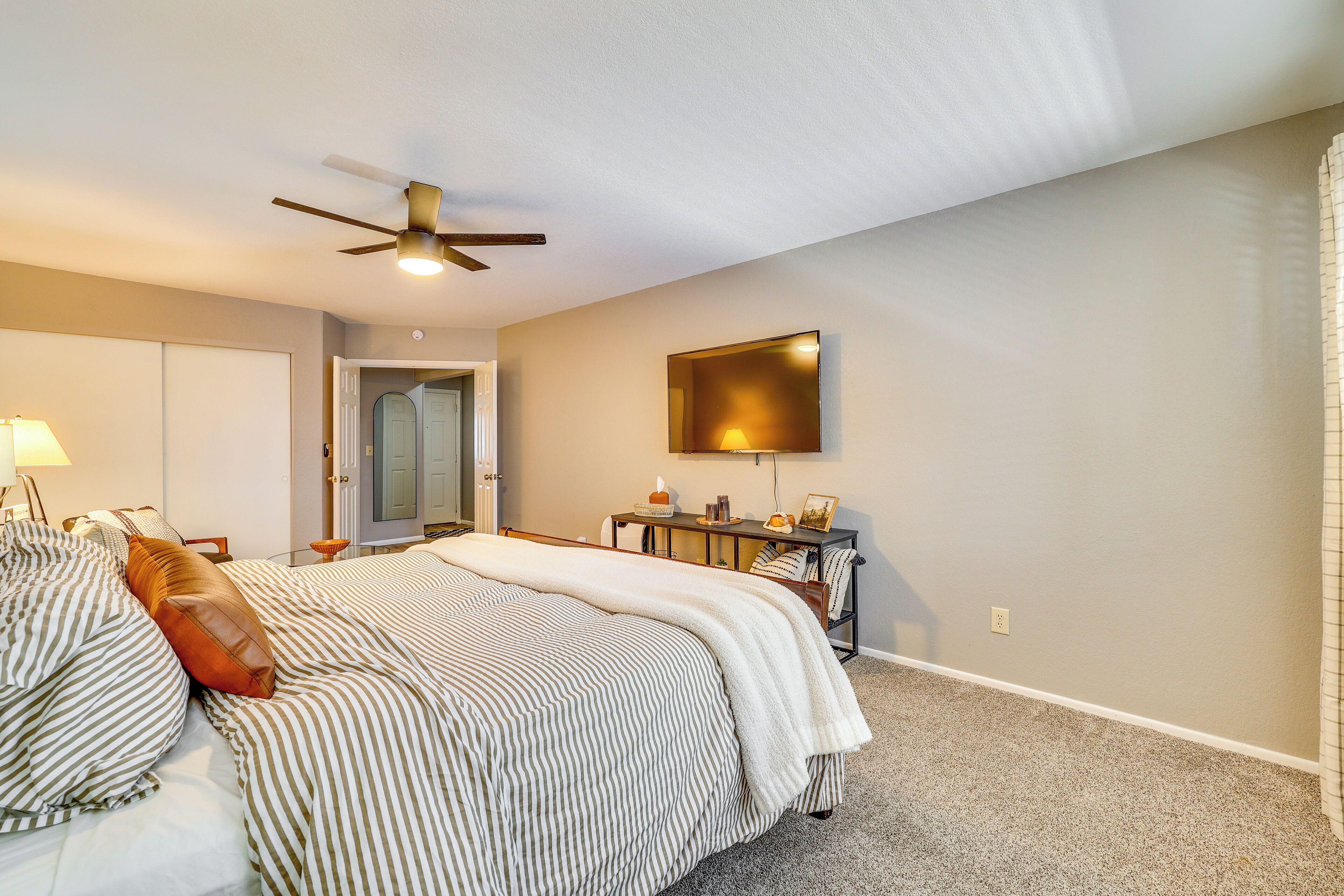 A bedroom with a bed, a ceiling fan, a bedside table with a lamp, and a door leading to another room.