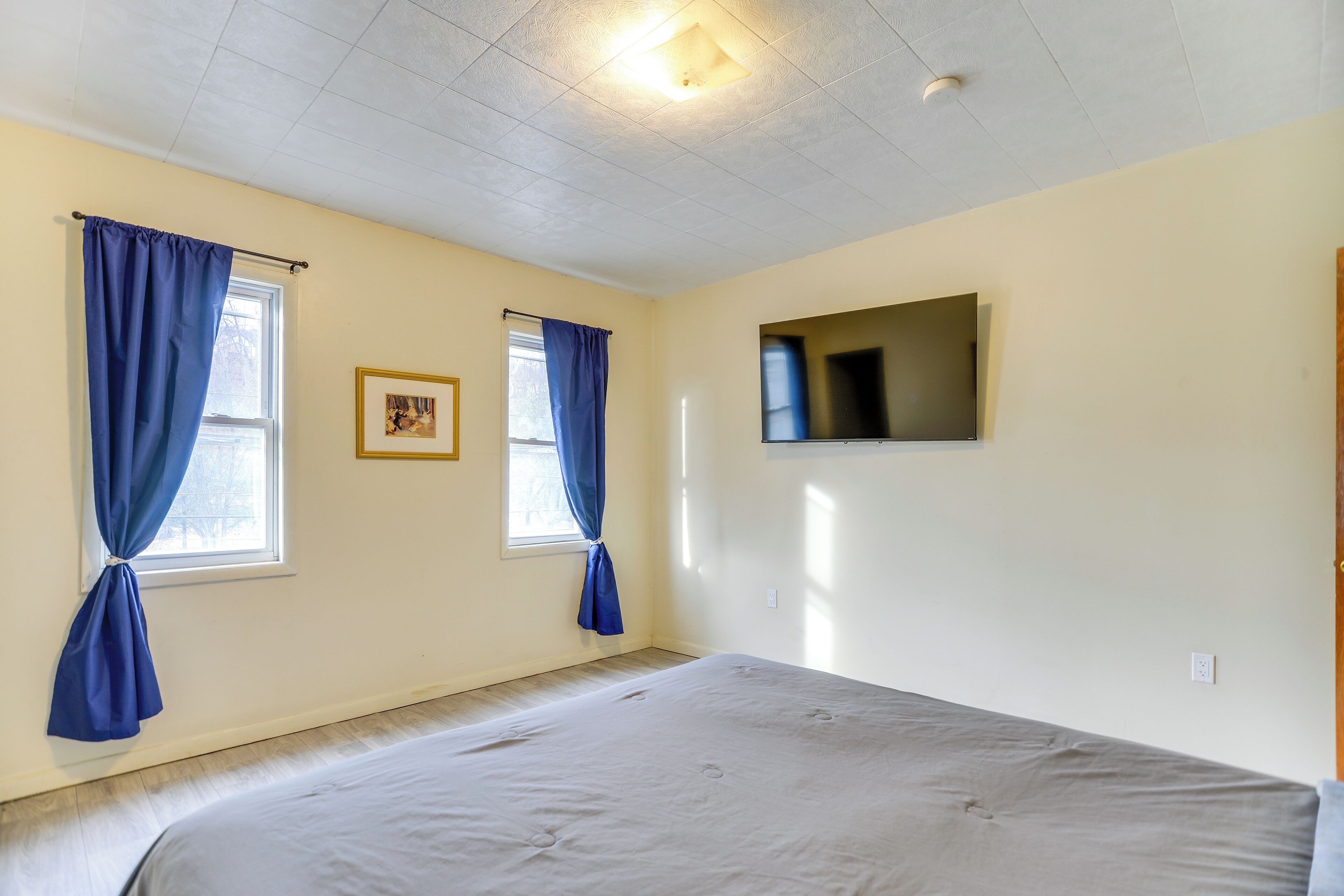 A bedroom with a bed, two windows with blue curtains, a flat-screen TV mounted on the wall, and a ceiling light fixture.