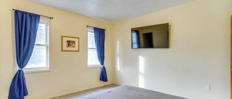 A bedroom with a bed, two windows with blue curtains, a flat-screen TV mounted on the wall, and a ceiling light fixture.