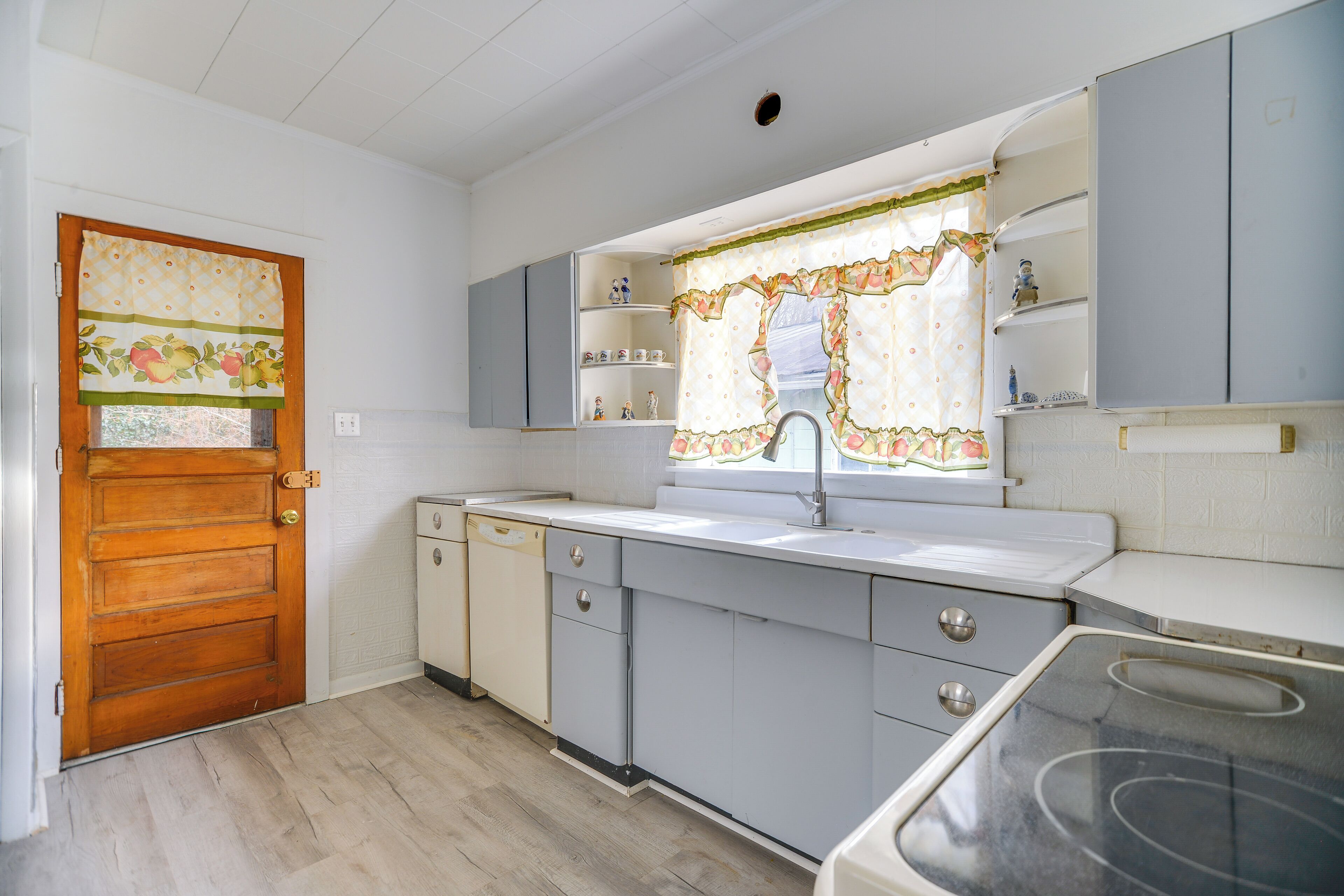A kitchen with a wooden door, a window with a floral curtain, and light-colored cabinets.