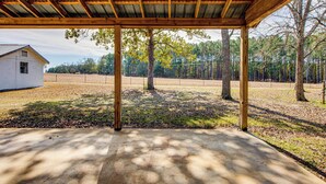 A covered outdoor area with a concrete floor, wooden beams, and a view of a house and trees in the background. - Cozy Barn Apt w/ 5-acre Pond in South Georgia! (Doerun)