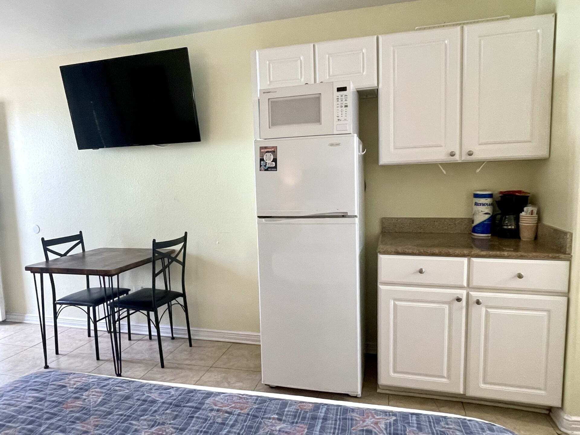 A compact kitchen with white cabinets, a white refrigerator, a microwave, and a small countertop with a coffee maker and can. There is a dining table with two chairs and a TV mounted on the wall.