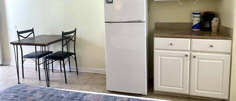 A compact kitchen with white cabinets, a white refrigerator, a microwave, and a small countertop with a coffee maker and can. There is a dining table with two chairs and a TV mounted on the wall.