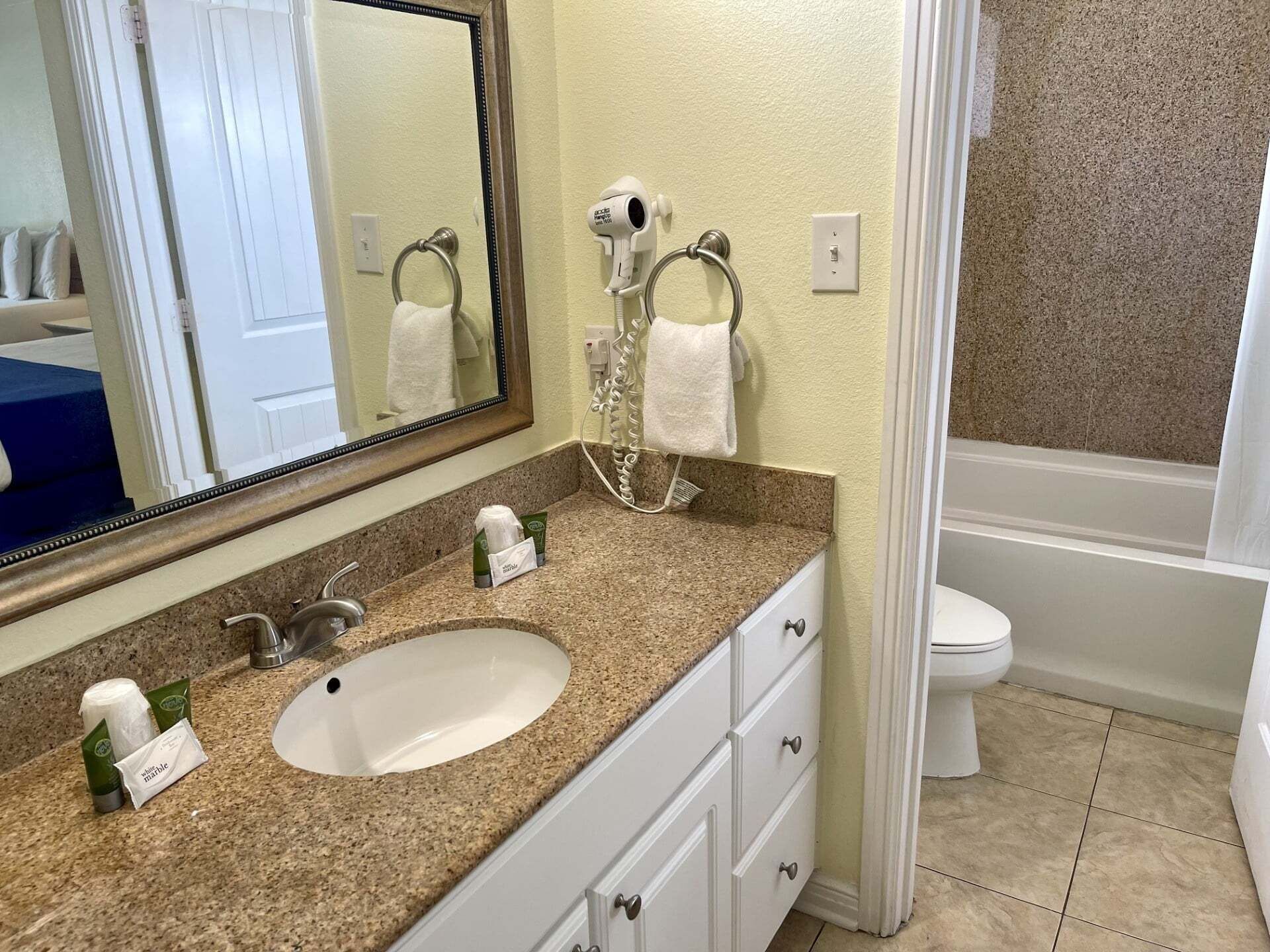 A bathroom with a granite countertop, white sink, and a large mirror. There is a hairdryer on the wall, a towel ring, and a toilet visible.