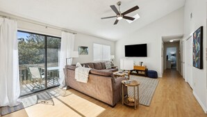 A spacious living room with a large sofa, a ceiling fan, a flat-screen TV, and a balcony with a sliding glass door. - Forest Ridge U8 2 Bedroom Condo (Fernandina Beach)