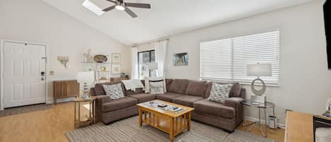 A living room with a brown sofa set, a wooden coffee table, a ceiling fan, and a large window with blinds.