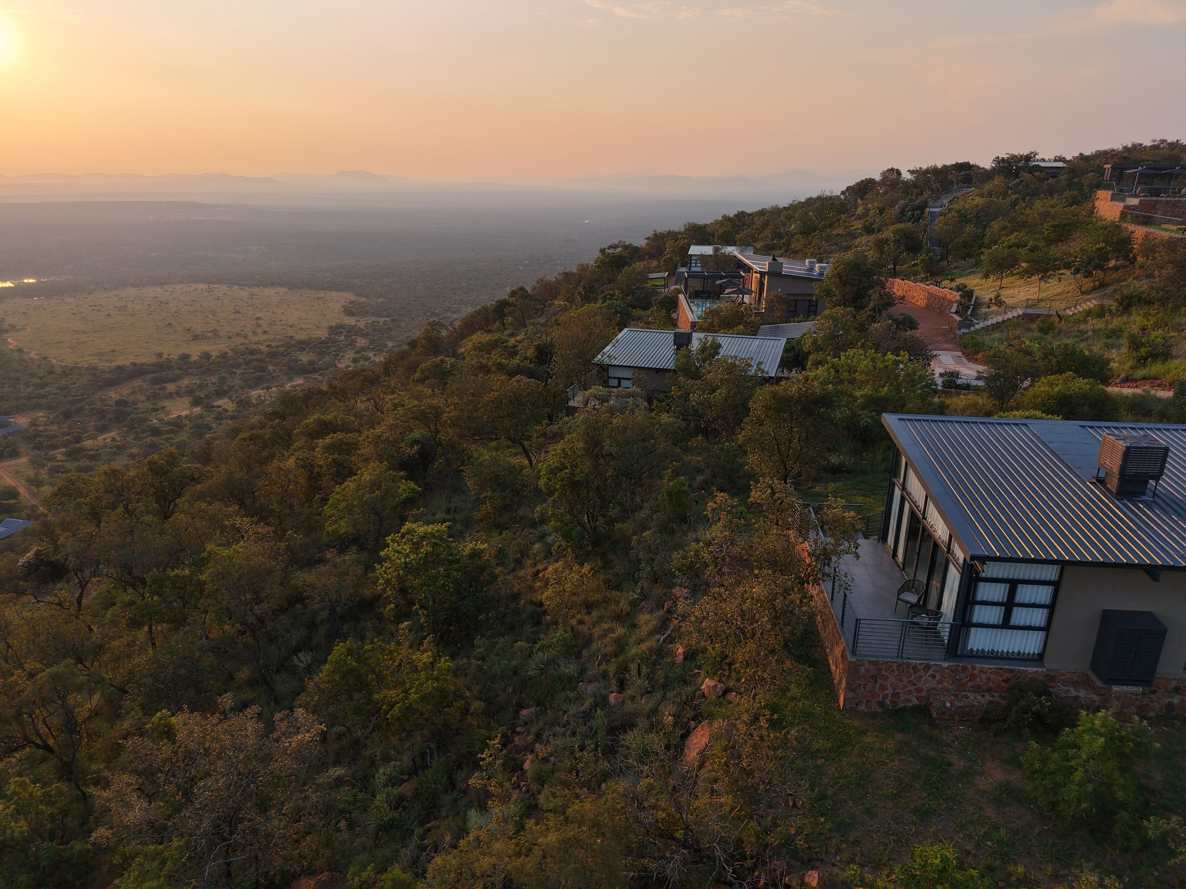 A hillside property with multiple buildings, including a house with a stone facade and a modern design, surrounded by lush greenery and a view of the landscape.