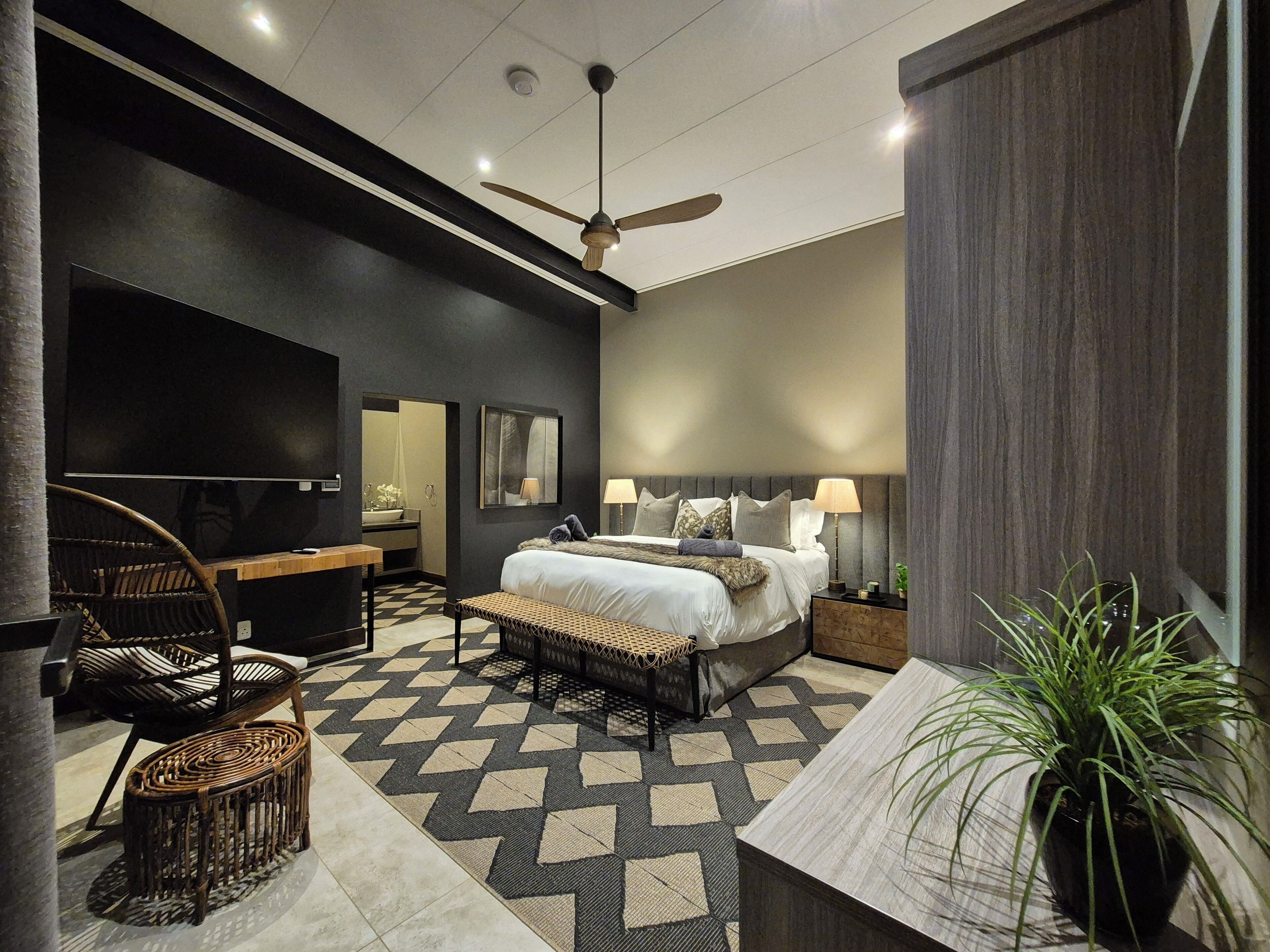 Quarto de hotel moderno com uma cama grande, ventilador de teto, televisão, escrivaninha, cadeira, planta em vaso e piso estampado.