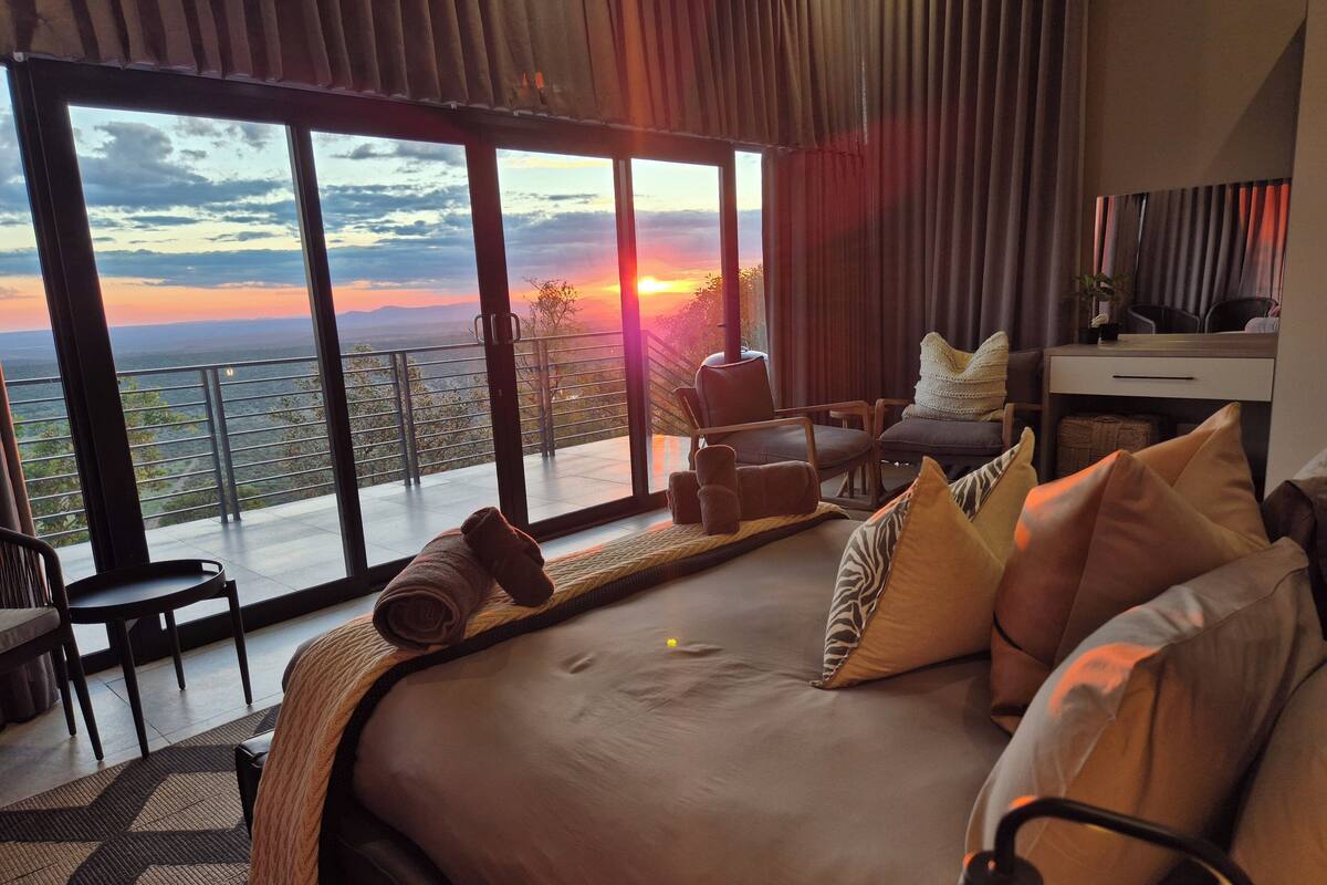 Une chambre avec un grand lit, un bureau et une chaise. De grandes fenêtres offrent une vue sur le coucher du soleil.