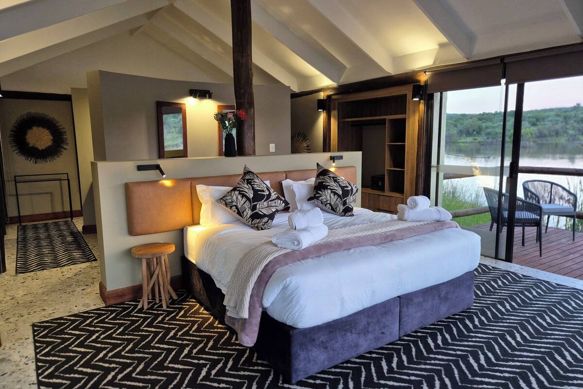 Une chambre avec un grand lit, une table de chevet, une lampe, une vue sur l’extérieur et un plafond en bois.