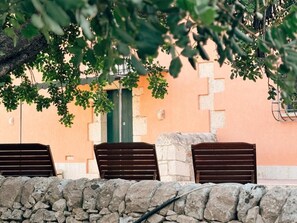 A stone wall with three wooden chairs facing a building with a green door and a peach-colored wall.