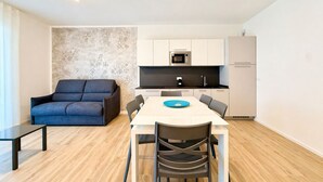 A modern kitchen with white cabinets, a microwave, and a refrigerator. There is a dining table with chairs and a blue plate. A dark sofa is against the wall. - Aurora Sand 2 - Carraro immobiliare Jesolo (Lido di Jesolo)