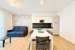 Eine moderne Küche mit weißen Schränken, einer Mikrowelle und einem Kühlschrank. Ein Esstisch mit Stühlen und einem blauen Teller sind vorhanden. Gegenüber der Wand steht ein dunkles Sofa.