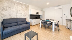 A modern living room with a sofa, a dining table with chairs, a kitchen area with cabinets and appliances, and a decorative wall. - Aurora Sand 2 - Carraro immobiliare Jesolo (Lido di Jesolo)