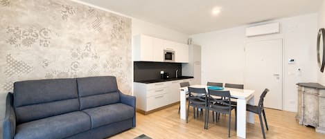 A modern living room with a sofa, a dining table with chairs, a kitchen area with cabinets and appliances, and a decorative wall.