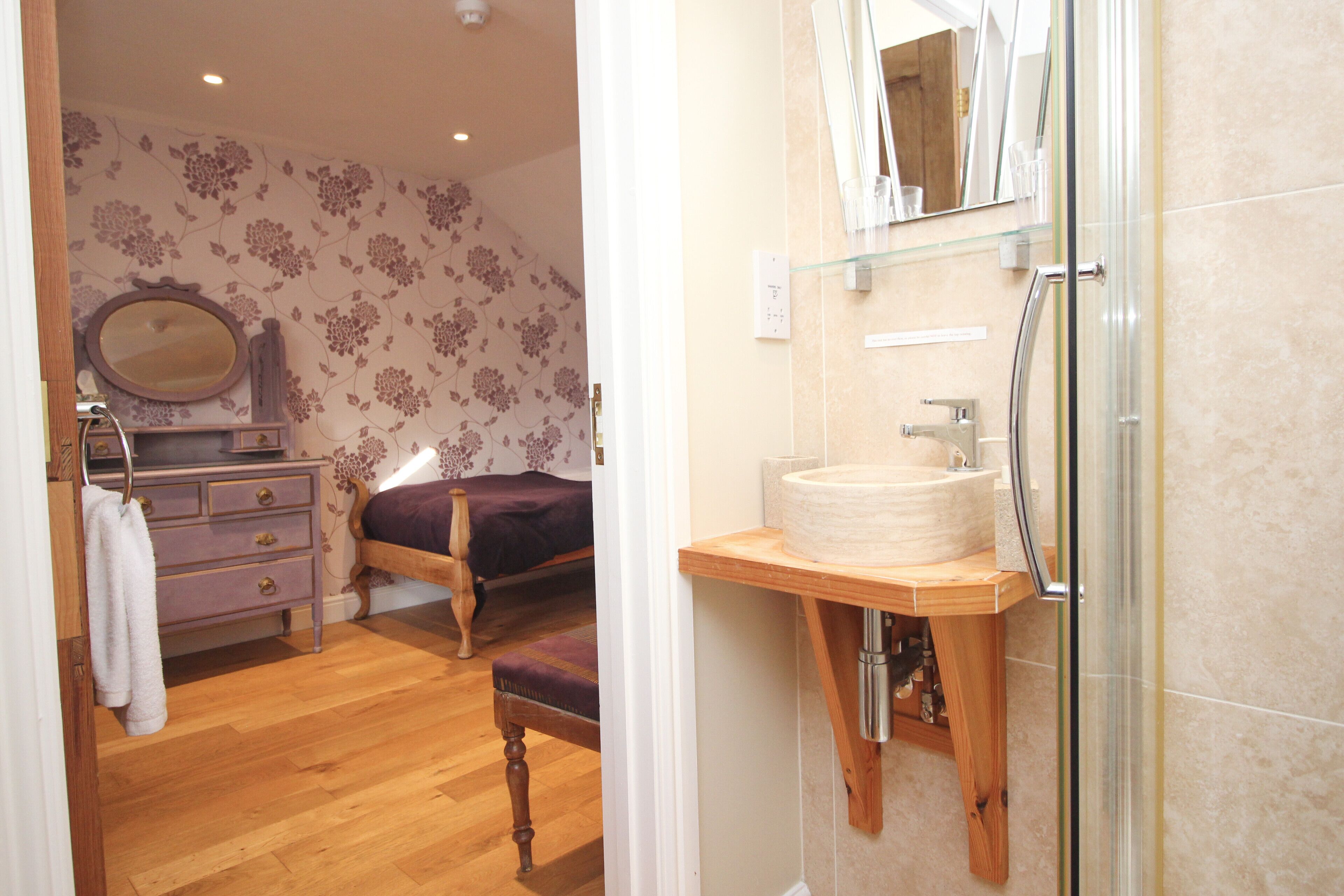A bedroom with a wooden floor, a bed with a purple bedspread, a dresser with a mirror, and a bathroom with a glass shower enclosure and a sink.