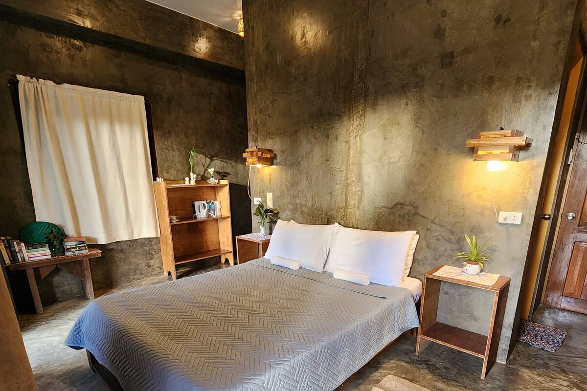 Un dormitorio con una cama grande, una mesita de noche de madera, una pequeña mesa y una estantería con libros y objetos decorativos.