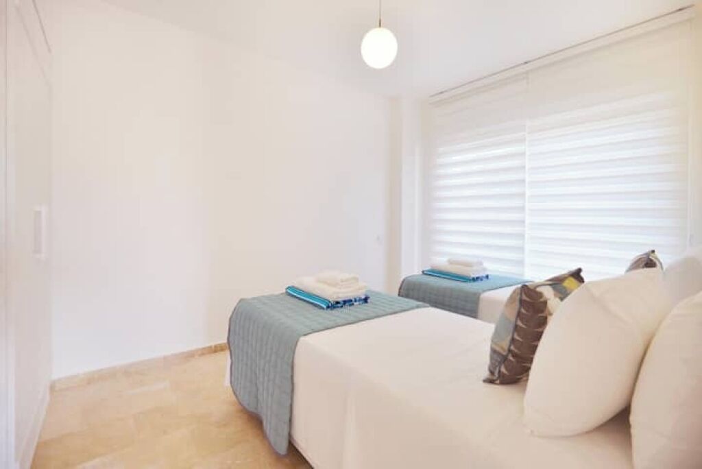 Une chambre d’hôtel avec deux lits, un canapé, une fenêtre avec des stores et un luminaire suspendu.
