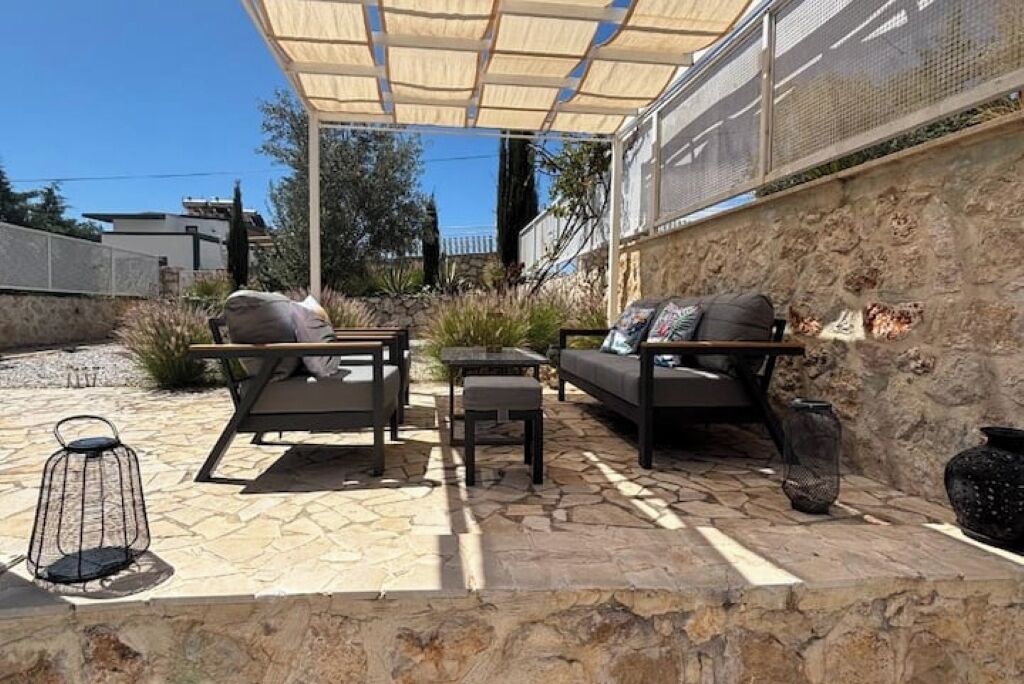 Un patio al aire libre con suelo de piedra, una mesa de madera, dos sofás y una pérgola que da sombra.