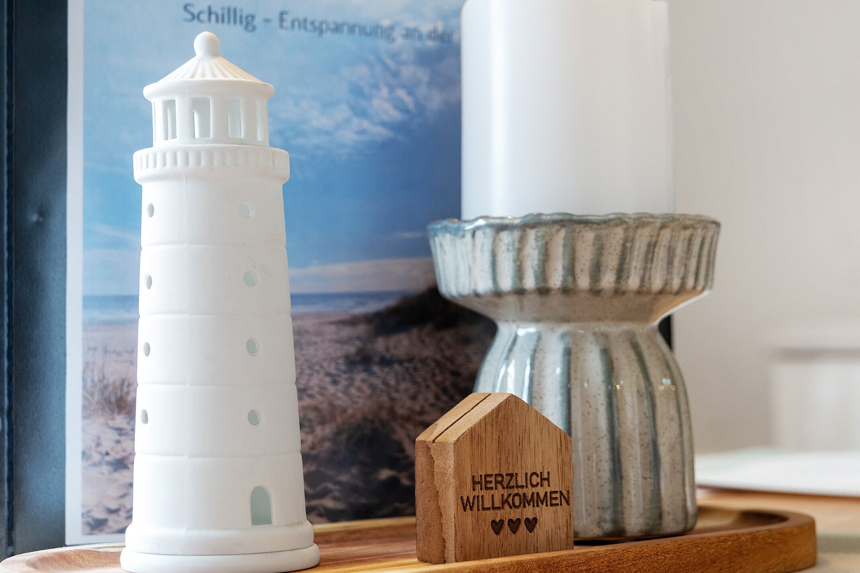 Een witte vuurtoren, een houten bord met 'HERZLICH WELCOME' en een keramieken kaarsenhoudertje op een houten dienblad.