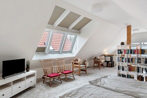 A bedroom with a sloped ceiling, a TV, two wooden chairs with cushions, a bookshelf, and a window with a view of red-roofed buildings. - Holiday home 'Me(e)rzeit' in Schillig (Schillig)