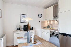 A modern kitchen with white cabinetry, a coffee maker, a clock, and a dining area with a table and chairs. - Holiday Home Me(e)rzeit in Schillig – Close to the Beach (Schillig)