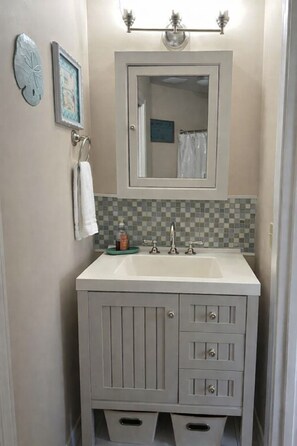 A bathroom with a vanity, sink, and mirror. There are two towel rings on the wall, a small bottle on the sink, and a tiled backsplash. - Peek of the Gulf - Cozy Beach Studio (Panama City Beach)