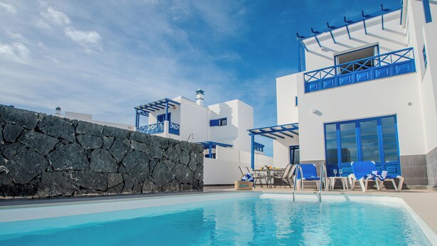 A modern pool area with a stone wall, lounge chairs, and a clear blue sky. - Villa Vereda Del Sur - Sea View, Pool, close to promenade, shops and supermarket (Playa Blanca)