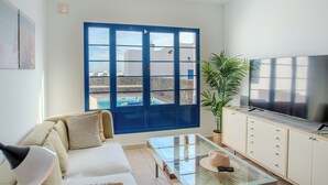 A modern living room with a sofa, a glass coffee table, a television, and a large window with a view of a swimming pool. - Villa Vereda Del Sur - Sea View, Pool, close to promenade, shops and supermarket (Playa Blanca)