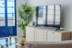 Ein modernes Wohnzimmer mit einem Flachbildfernseher auf einer Holzkommode, einer großen Topfpflanze und einem Glastisch mit Hut und Sonnenbrille.