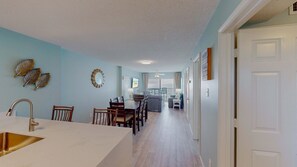 A modern kitchen with a white countertop and stainless steel sink, a dining area with wooden chairs and a table, and a living room with a sofa and a TV. - 2br/2ba Beachfront Condo With Stunning Ocean Views + Heated Indoor/outdoor Pool (Orange Beach)