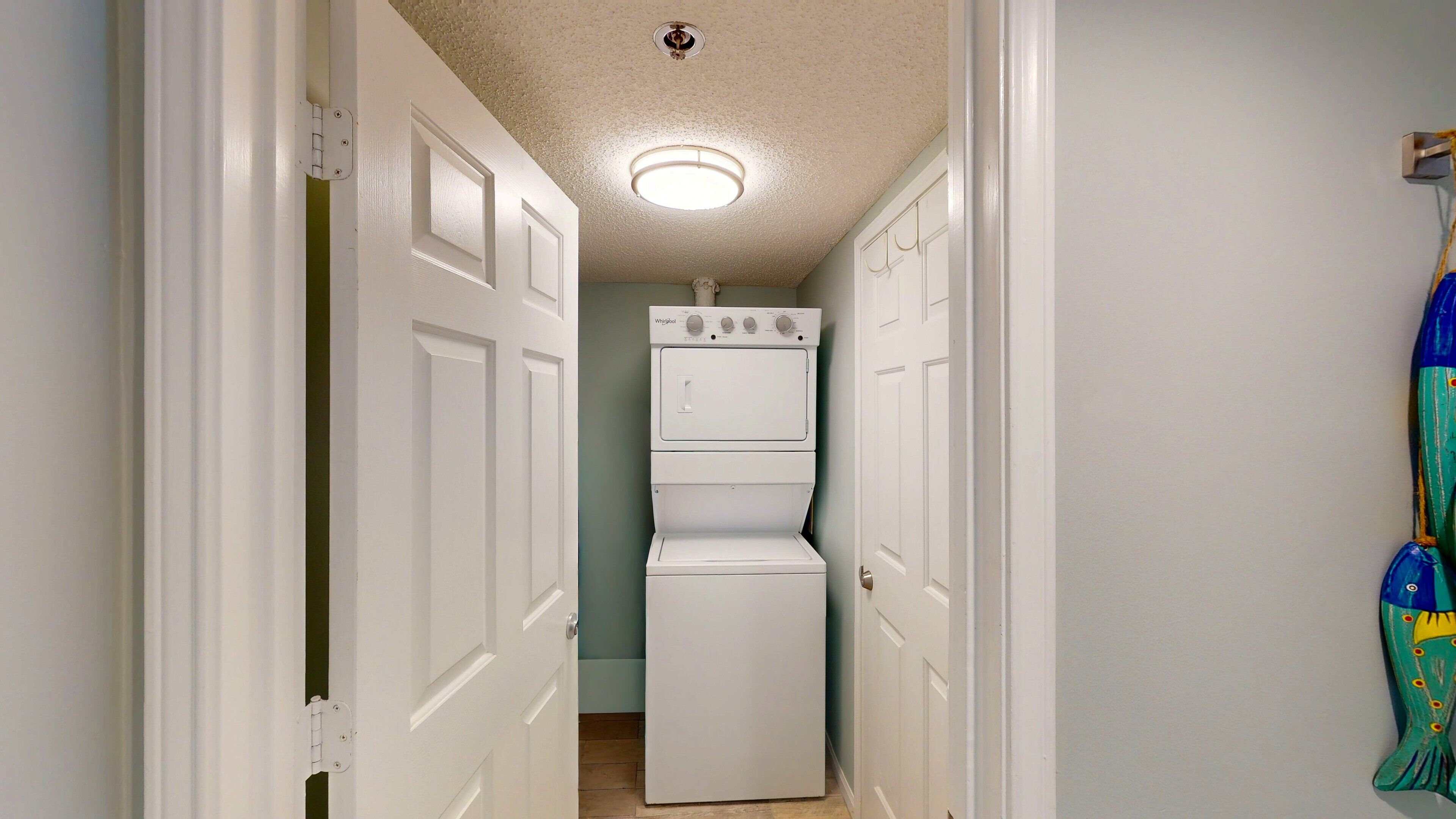 A laundry room with a stacked washer and dryer, a ceiling light, and a door leading to another room.