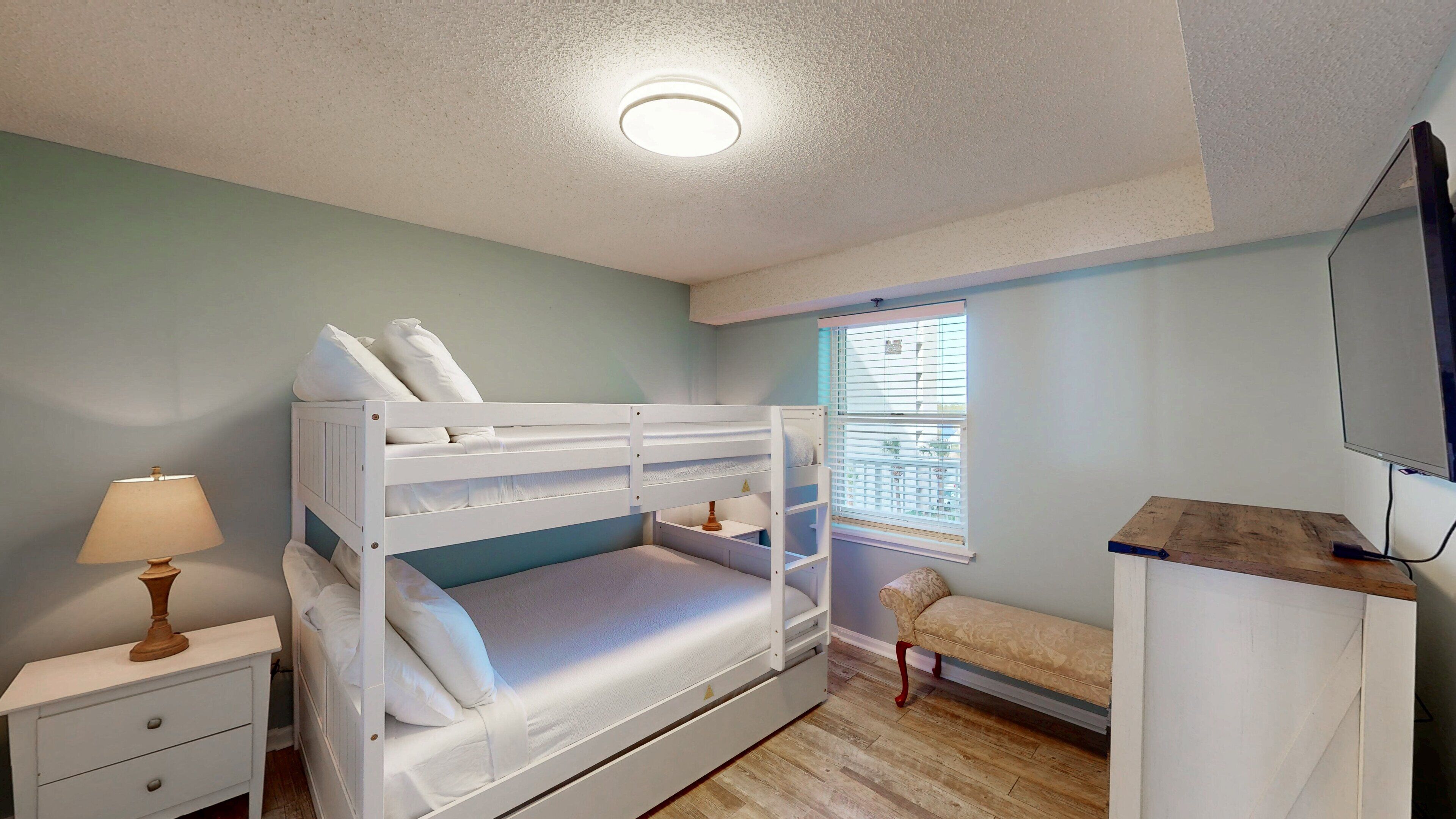 A bedroom with bunk beds, a bedside table with a lamp, a window with blinds, a sofa, and a TV mounted on the wall.