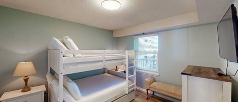A bedroom with bunk beds, a bedside table with a lamp, a window with blinds, a sofa, and a TV mounted on the wall.