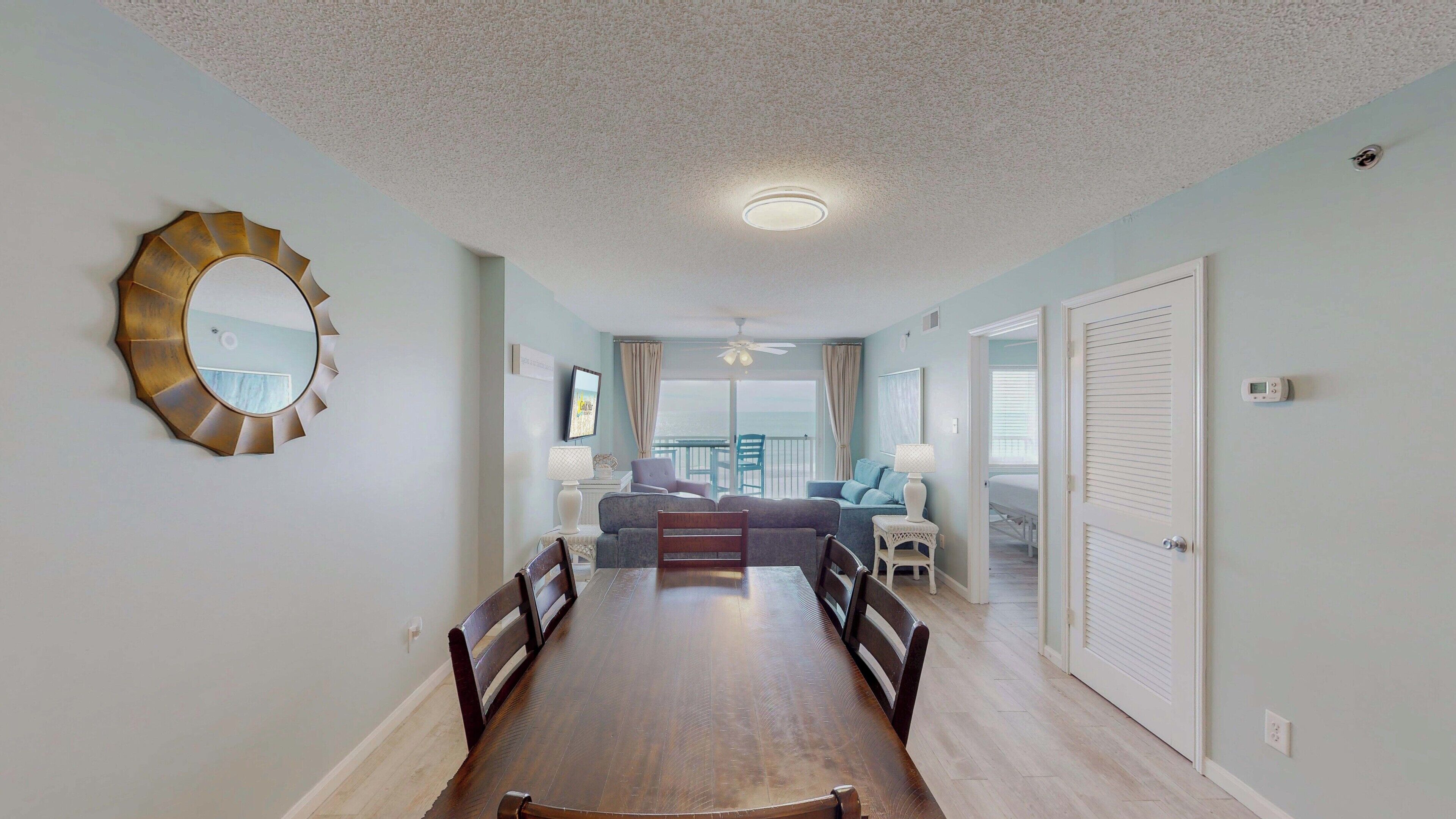A dining area with a wooden table and chairs, a round mirror, and a view of the ocean through a sliding glass door.